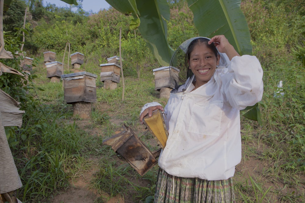 A smiling Guatemalan woman in a bee suit poses with her beehivevs.