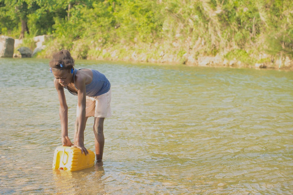 A young girl gathers water from a stream into a yellow cannister.