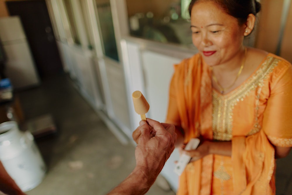 A nepali woman in bright orange kurta suruwal hands an ice cream to a customer off camera