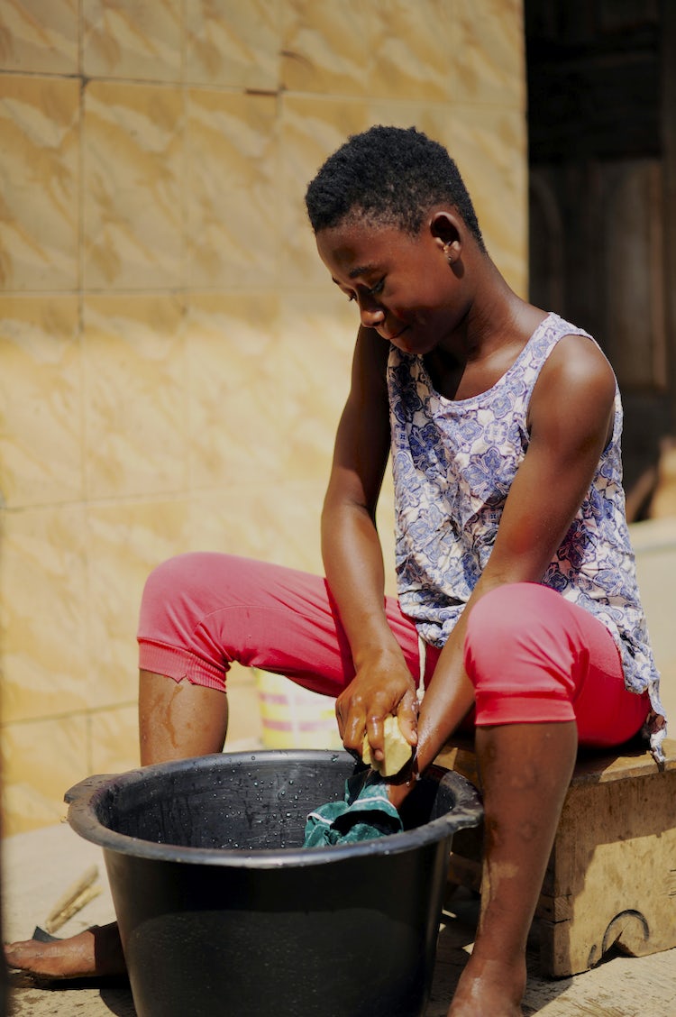 A Ghanese girl sits near a basin, washing her school uniform with the soap her mother made.