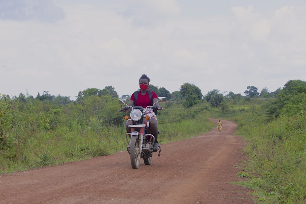 Leah Amongi rides her motorcycle on a smooth dirt road. She is wearing a red face mask, a red shirt and a red and white skirt.