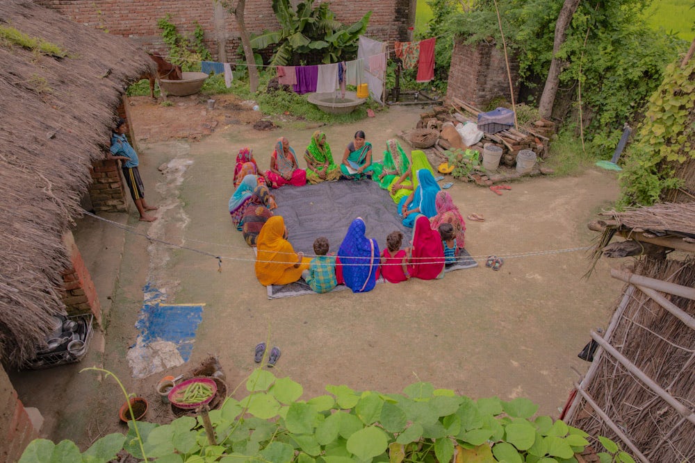 A group of women sitting in a circle in the lawn in front of a house in a village, and talking with each other.