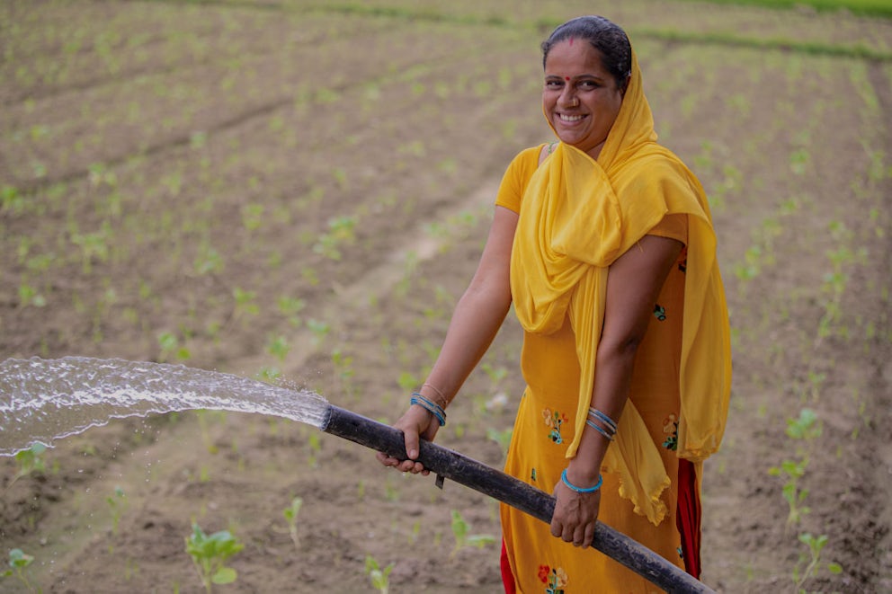 A Nepali woman wearing a bright yellow shawl waters her vegetables with a hose.