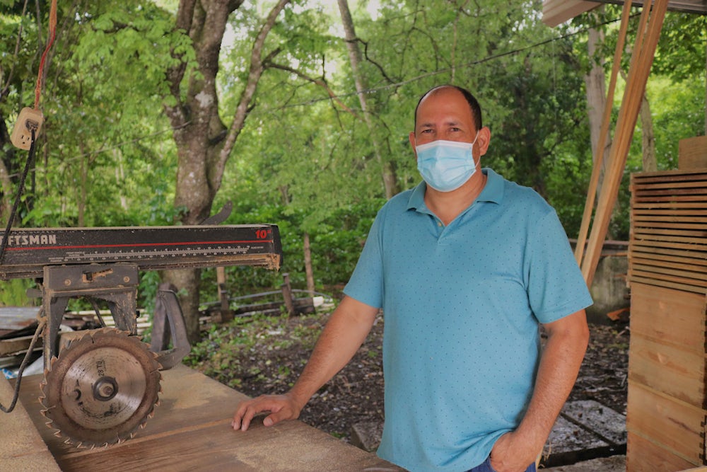 A Honduran man wearing a mask and blue shirt stands by his carpentry workshop.