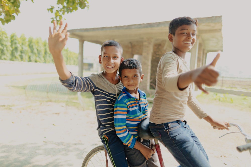 Three boys smile and wave while riding a bicycle.