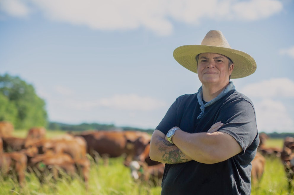 Donna Kilpatrick stands with her arms crossed in front of a herd of brown cattle.
