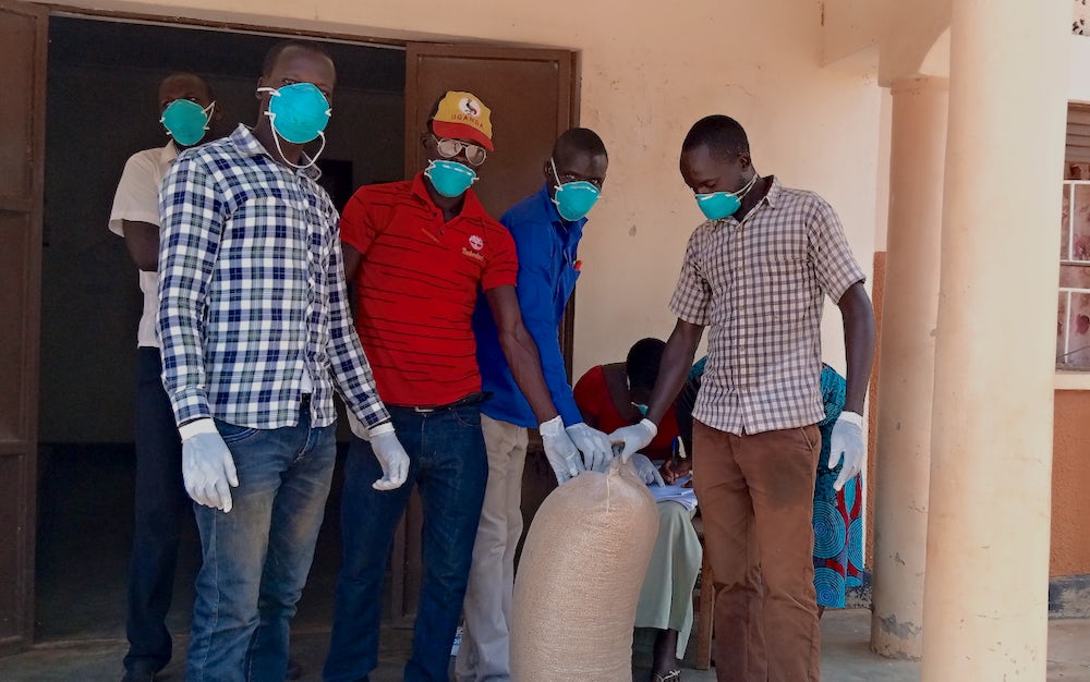 A group of men gather around a bag of seeds. All are wearing blue masks in accordance to cdc guidelines. 