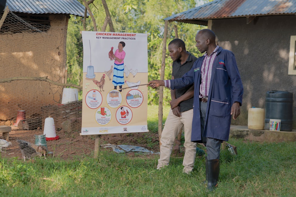 A community vaccinator gives a presentation on improved chicken management techniques to a poultry farmer standing beside him.
