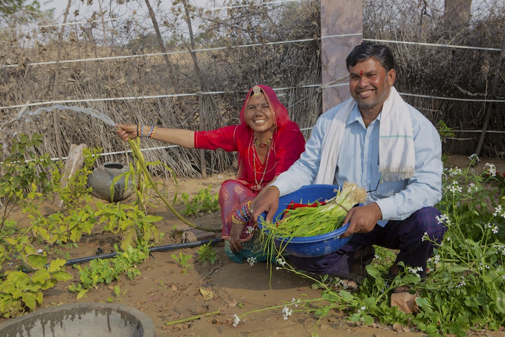 An Indian woman and man crouch down in their garden, smile at the camera and holding vegetables.