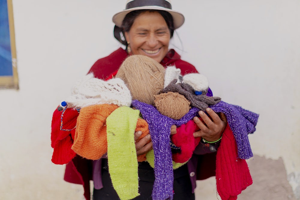 A woman in a bright red cloak stands holding an armfull of knitted goods and hand-spun yarns