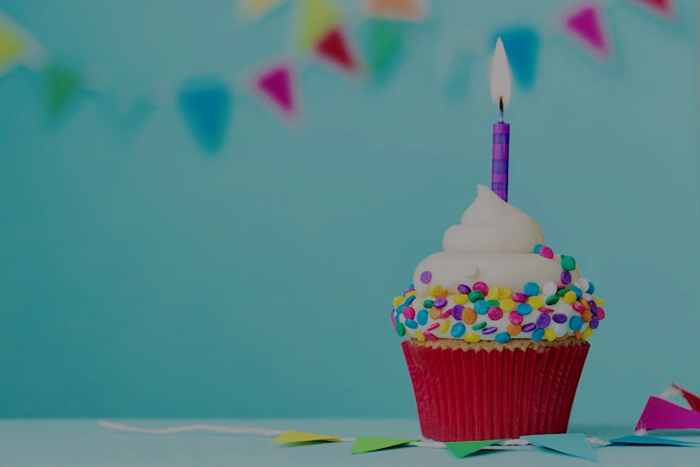 A festive cupcake with a lit birthday candle sits amongst party banners and a light blue background.